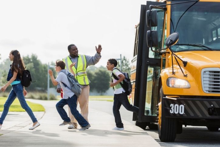 Children cross the street in front of a school bus. A crossing guard in a neon vest holds up his hand to direct traffic.